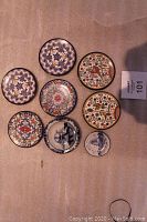 Top view of 7 small hand painted round plaques and plates arranged on a beige cloth, showing various Spanish folk art designs and a small Delft plate.
