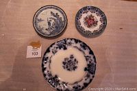 Three plates arranged on beige fabric: large blue and white plate with floral design, smaller floral multicolor plate, Asian-themed plate with metal ring backing.