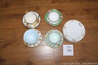 Five vintage tea cups and four hand-painted breakfast plates displayed on wooden floor.