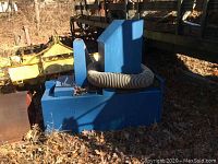Photo of blue seed blower machine with hose wrapped around the base, sitting outdoors among leaves.