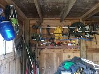 Photo inside a wooden garden shed showing various hanging and leaning yard tools including rakes, hand trimmers, forks, a blue plastic sprinkler and a blue hammer.
