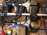 Close-up of two gas-powered weed wackers with white gas tanks and some hoses in background.