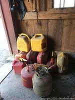 Photo showing a collection of yellow and red gas cans stacked against a wooden wall in a shed environment, along with a metallic propane tank and a red kerosene container.