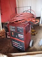 Battery charger with cord coiled on top sitting on a garage floor with a pegboard and buckets in background.