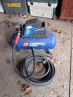 Full side view of the blue Campbell Hausi air compressor with attached black coiled hose on floor.