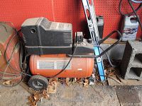 Side view of Craftsman orange air compressor with black motor housing and two wheels. Unit is on a dirty garage floor with leaves.