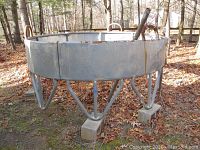 Full view of a large metal outdoor hay feeder showing the circular shape and metal support legs on concrete blocks in a wooded area.