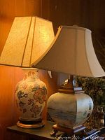 Two table lamps shown side by side on a table, with different designs on ceramic bases and fabric lampshades.