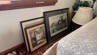 Three framed prints leaning against a wall near a window and bed. One shows a hunting scene, another a rustic barn, and the third a lighter framed print.
