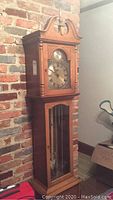 Full view of Ridgeway grandmother clock showing wooden case and decorative top finial next to brick wall.