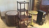 Photo shows three-tier wooden shelf with single drawer and octagonal wooden table with dark finish, heater and a small yellow side table in background.