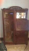 Front view of antique wooden secretary desk showing glass door cabinet on left with wooden frame and missing shelves, mirror above the drop-front writing area on right, and ornate carved wooden accents on top