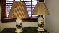 Two matching ceramic hunt scene table lamps, each with a different pleated lamp shade, shown side by side on a surface in front of window blinds.