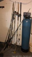 Overview of six fishing rods with PENN reels leaning against a white wall near blue tank