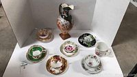 View of six tea cups with matching saucers and decorative ceramic pitcher arranged inside a white photo box on a concrete floor.