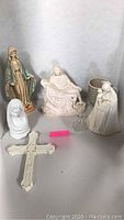 Religious figurines including painted Virgin Mary statue, white pietà Jesus and Mary figurine, unpainted Virgin Mary, M J Hummel figurine, small glass angel, and cream Lenox cross, arranged on table