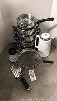 Photo of stainless steel cookware including saucepans, stockpots, mesh strainer, white floral thermos, cheese grater, and small ceramic container