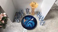 Overall lot with blue art dish in center, yellow-tan vase behind dish, and eight glasses arranged around