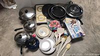 Photo of entire lot showing Revere Ware pots and pans, blue kettle, kitchen utensils, hot pads, plates, and box of plastic wrap on floor.