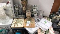 Image showing palm tree ceramic mugs, glass cup, wooden cutting boards, decorative tiles, tablecloth, napkins, and candle jar as part of the palm tree collection.