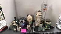 Group photo of assorted beer steins, mugs, bottle stoppers, and metal tools arranged on a black surface against white background.