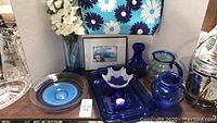 Photo showing cobalt blue glass items including baking dishes, vases, pitcher, candle holders, decorative bowl, and framed picture.