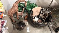 Overview photo of the lot showing giraffe sculptures of varying sizes, woven basket with towels, faux plants, and a dark vase on the floor.