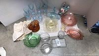 Wide angle photo showing assortment of Depression glass pieces in various colors, doilies, and decorative swan figurines on grey floor against white wall