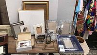 Photo showing multiple glass and metal picture frames in different sizes and styles arranged on a table, with easel stands and plain wooden frames in background.