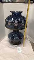 Full side view of large antique blue glass lamp showing floral design and ornate metal base. Measuring tape shows height about 26 inches.