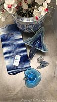 Photo showing large blue and white ceramic planter pot with white and pink artificial flowers, two blue glass swan figurines, Murano-style blue glass flower, and blue and white Afghan throw on floor.