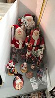 Multiple traditional Santa Claus figurines, including a Lenox Santa figurine with a toy bag, and several Santa-themed drinking glasses placed inside a box with a measuring tape showing size.
