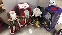 Two Santa Claus figurines with their original boxes placed behind them on a concrete floor.
