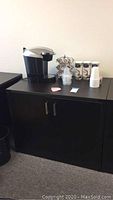 Wide view showing black office cabinet with closed two doors, silver handles, black Keurig coffee machine on top, along with K-Cup holder rack, canisters, and disposable cups.