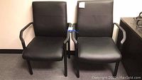 Photo showing two black office chairs side by side with blue tape on the armrest joint area, placed on office carpet.