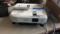 Full view of Epson projector with remote control resting on top. Projector is white with vent grills and lens visible.