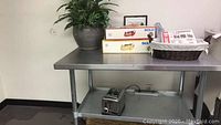 Metal kitchen office table with silk plant, kitchen supplies boxes, wicker basket, and toaster on lower shelf.