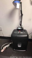 Photo of the black adjustable desk lamp placed on top of the black Staples paper shredder, showing whole items against white wall and black surface.