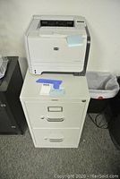 Front view of white HON two-drawer file cabinet with HP LaserJet printer on top, showing cabinet handles and lock