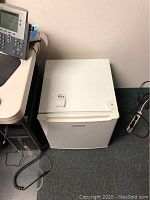 White Emerson mini refrigerator with front door closed, placed on grey carpet next to desk and power strip.