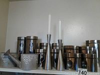 Full view of two decorative metal watering cans with floral patterns, eleven stainless steel storage containers arranged behind, and two stainless steel candle holders with white taper candles in front.