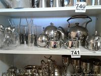 Shelf display of metal teapots, cream and sugar set, metal and glass beverage glasses, and tea strainers