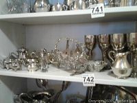 Shelf displaying various silverplated tea and coffee serving items including teaspoons, creamers, sugar bowls, and lidded containers.