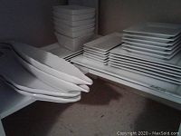 Multiple rectangular white china serving platters and trays stacked, along with small square dishes and bowls, white finish.