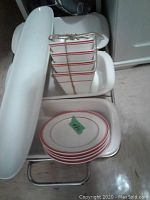 Set of white Corningware plates with red rim and white small square dishes tied with twine on a three-place metal serving dish holder, plus a long white dish next to it.