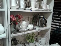 Shelves showing white ceramic cups and saucers, white plates, tea pot, flower arrangements, and a creamer and sugar bowl.