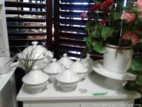 White ceramic tureen with ladle, six lidded soup bowls, ceramic cake stand, and planter with faux plant on a table in front of blinds.