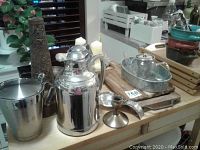 Photo showing stainless steel beverage container, stainless steel bucket, candle holders including tall decorative metal one and white candles, wooden trays, glass teapot, and wooden cutting board on tabletop.