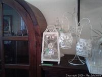Three white metal candle holders placed on a wooden sideboard: one large lantern style and two smaller hanging globe holders with white floral metalwork