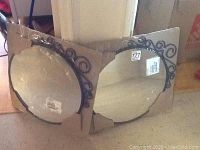 Two packaged round metal framed mirrors with decorative scroll designs, leaning against a wall.